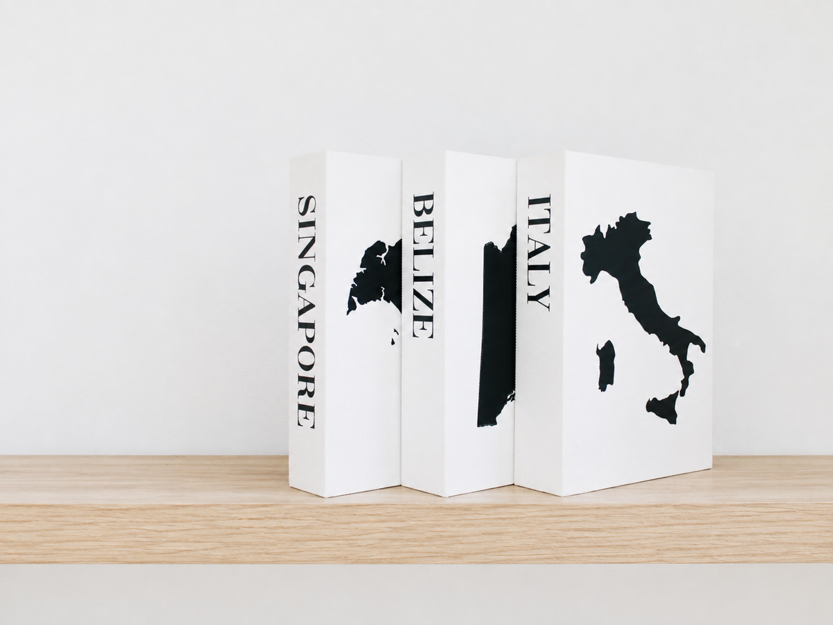 Three white decorative bookends shaped like book spines, featuring black map silhouettes and country names: Singapore, Belize, and Italy, displayed on a light wood surface against a white wall. Travel keepsake boxes to hold travel souvenirs in a decorative way. Faux book travel box. 