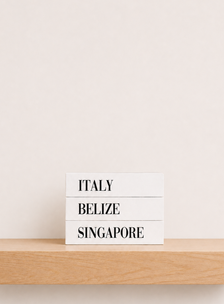 Three white rectangular blocks stacked on a light wood floating shelf, printed with the words 'ITALY', 'BELIZE', and 'SINGAPORE' in black serif text against a warm off-white wall. Travel keepsake boxes that look like bookshelf books but actually open like a box.